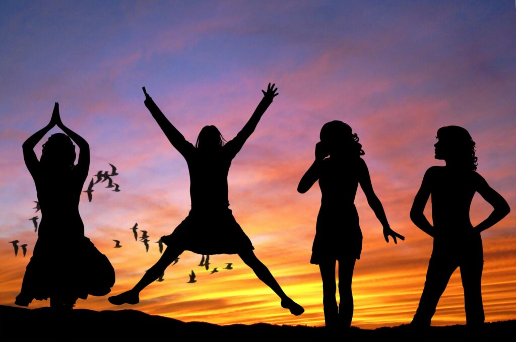 Silhouetted group of women joyfully jumping and posing against a vibrant sunset sky with birds flying.